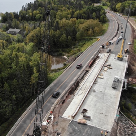 An aerial view of a road construction site next to a completed road, with a crane and construction equipment visible, surrounded by a forested area.