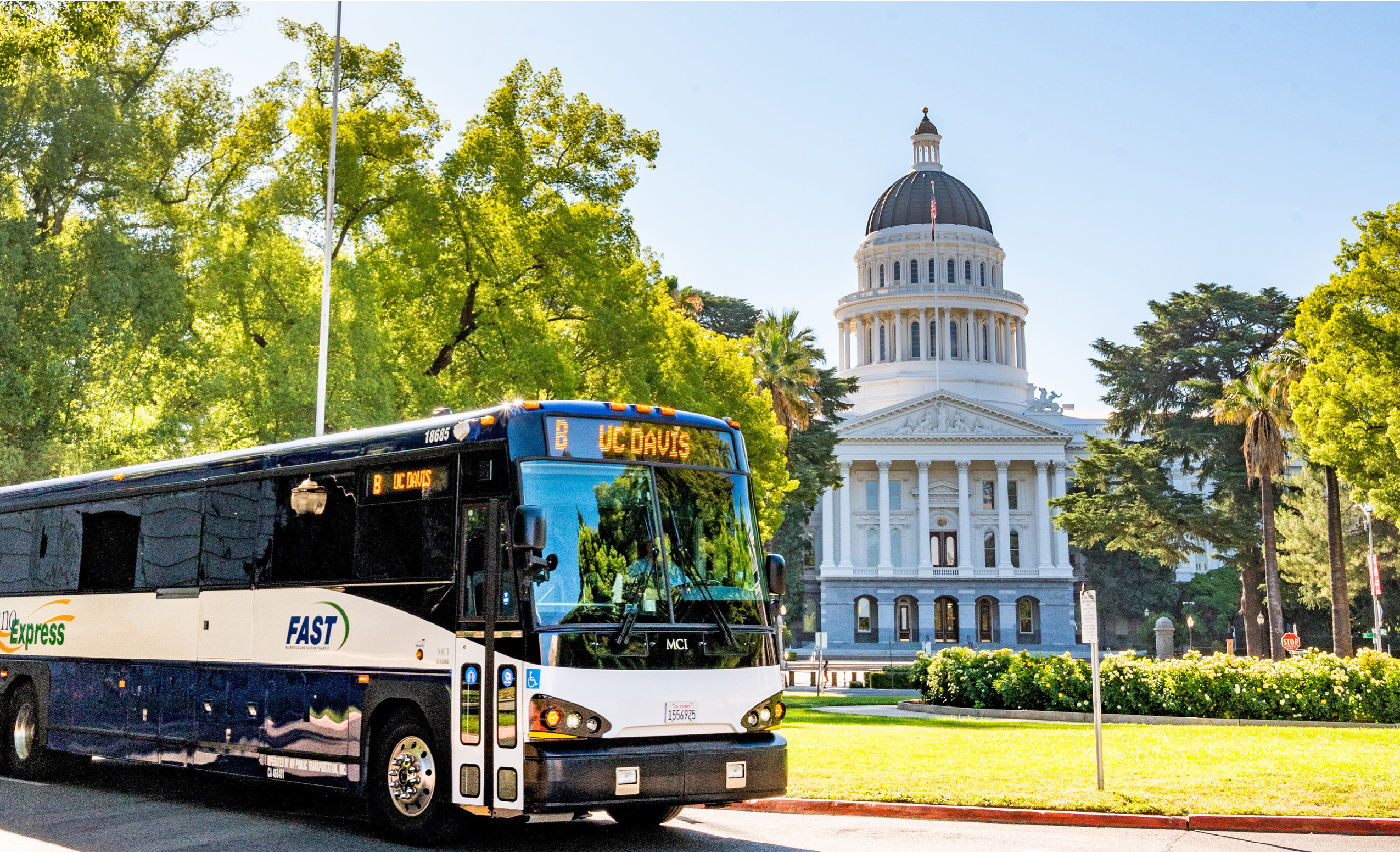 A bus labeled "UC Davis" from FAST public transit is driving in front of the California State Capitol building on a sunny day, surrounded by green trees and manicured landscaping.