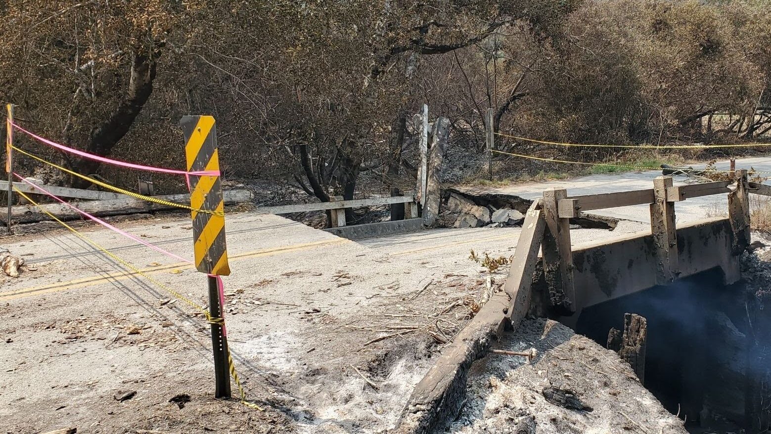 The image shows a damaged road and bridge in Santa Cruz County, with caution tape, a warning post, and evidence of fire damage, such as burnt trees and charred surroundings.