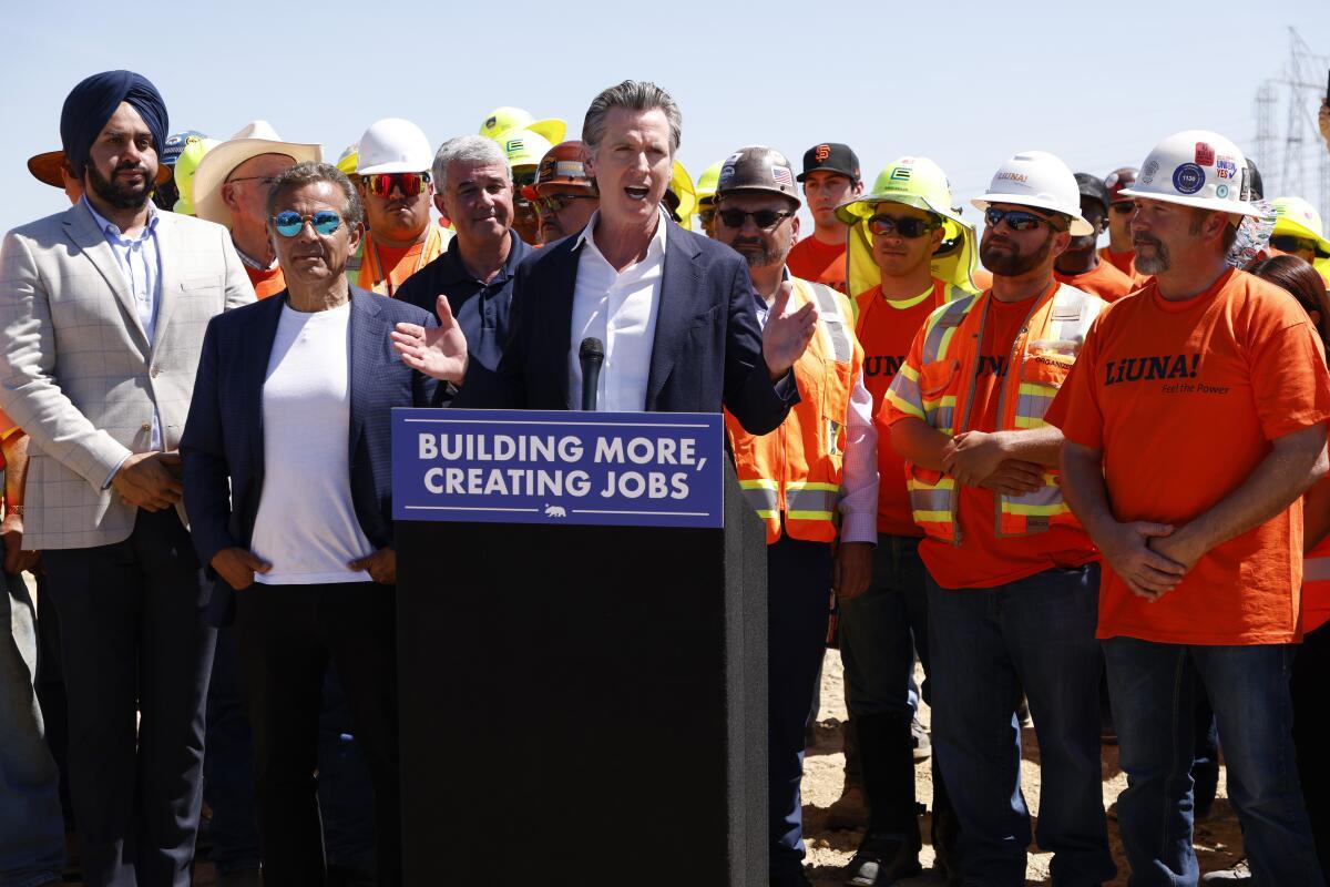 California Governor Gavin Newsom speaking at a podium labeled "Building More, Creating Jobs," surrounded by construction workers and officials at a job site.